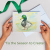 Card with a duck illustration held by hands with a green ribbon and envelope on a light background.