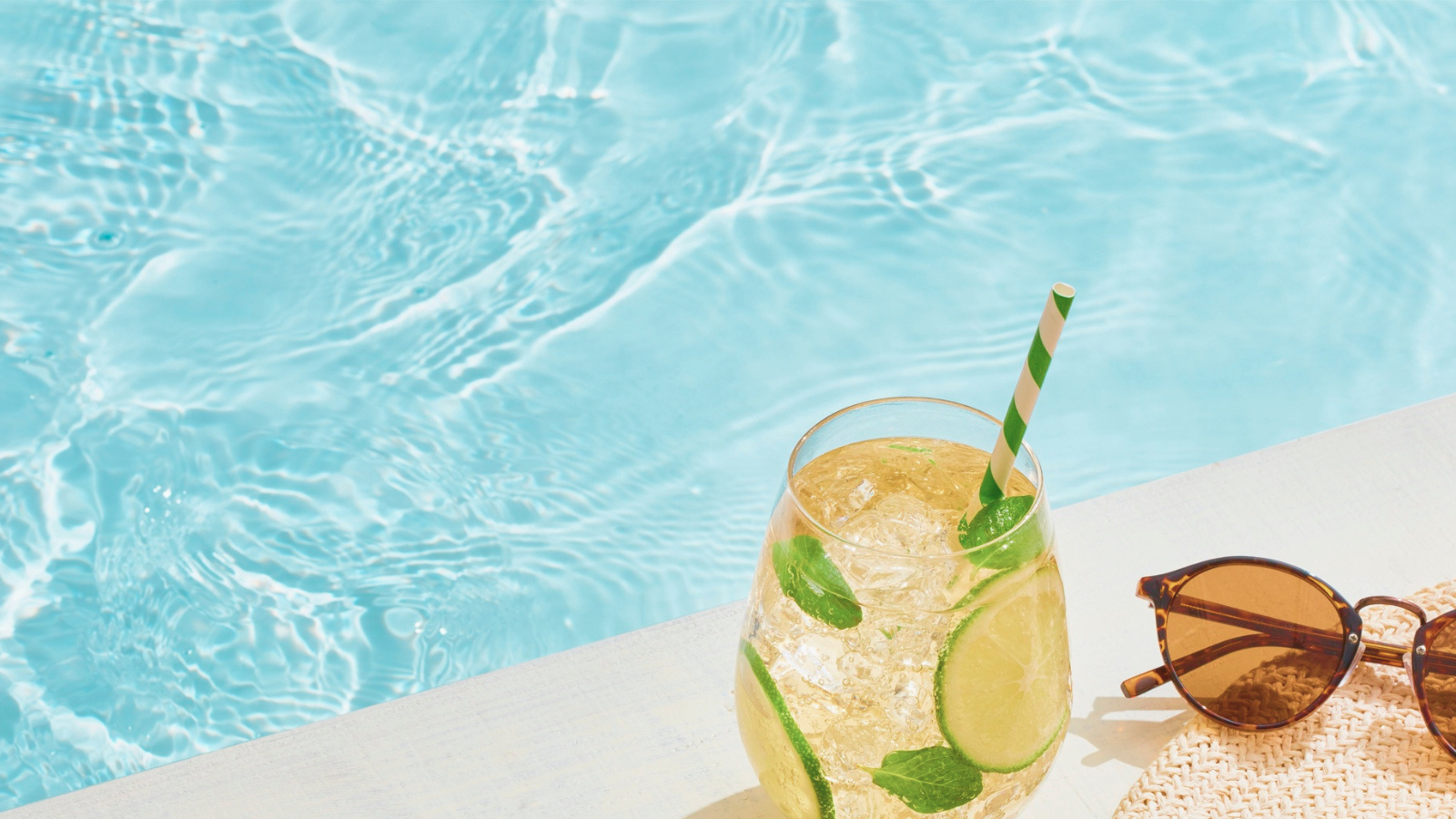Glass of lemonade with mint leaves and a straw, sunglasses, and a towel by a pool.